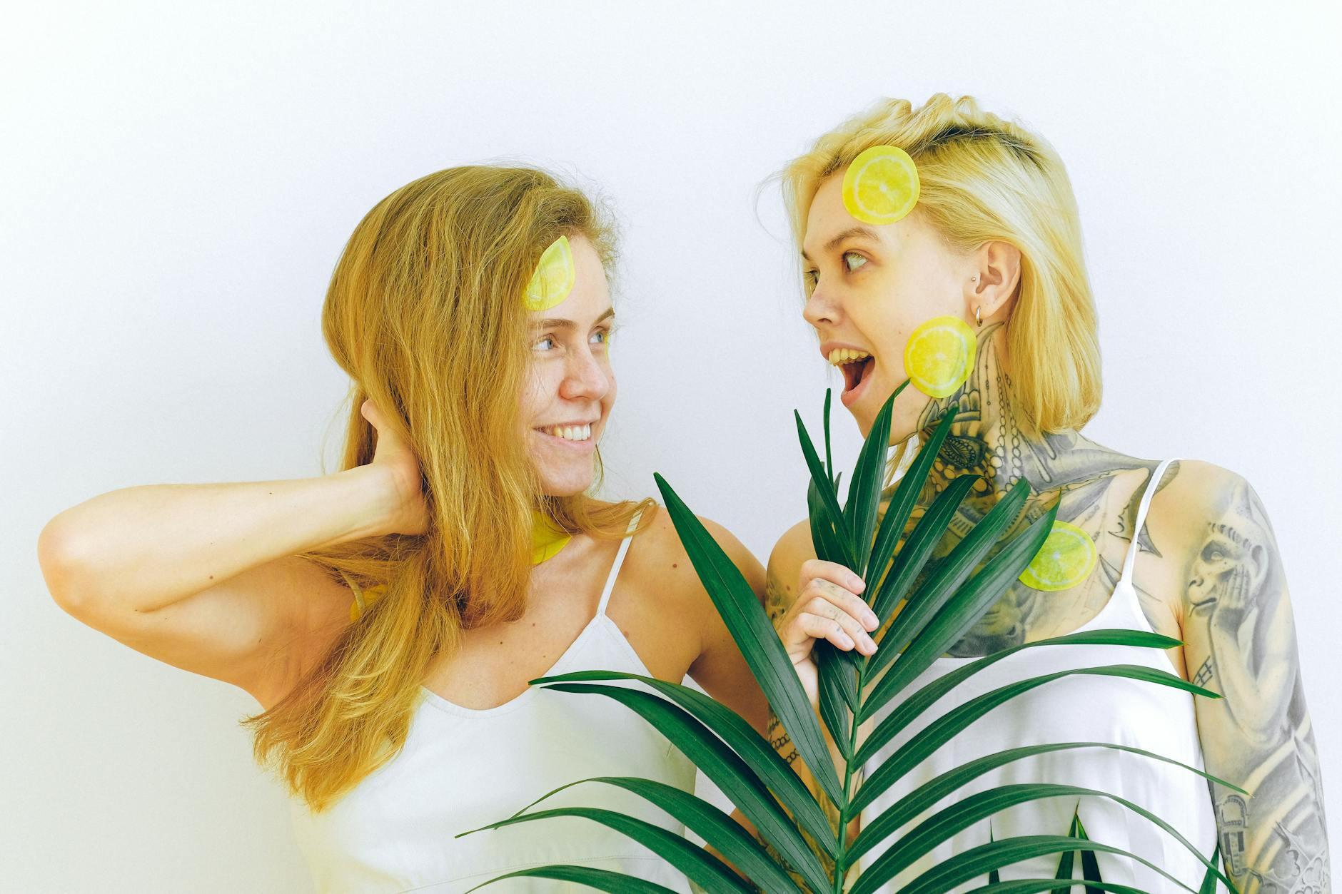 Two smiling women with lemon slices and tropical plant, expressing joy and fun indoors.