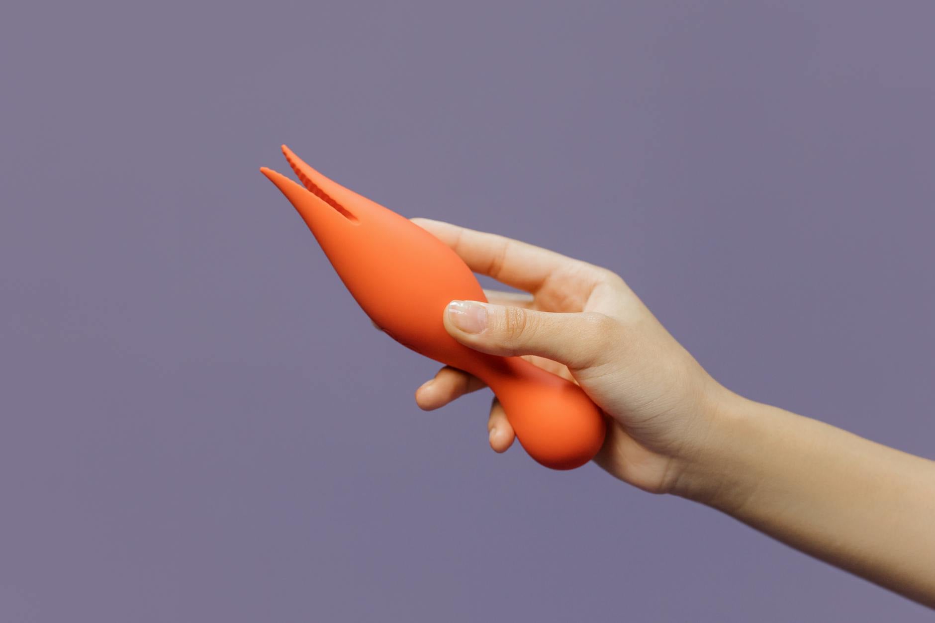 Close-up of a hand holding a citrus-colored clitoral vibrator against a minimalistic purple background, symbolizing the gentle return to sensual touch.