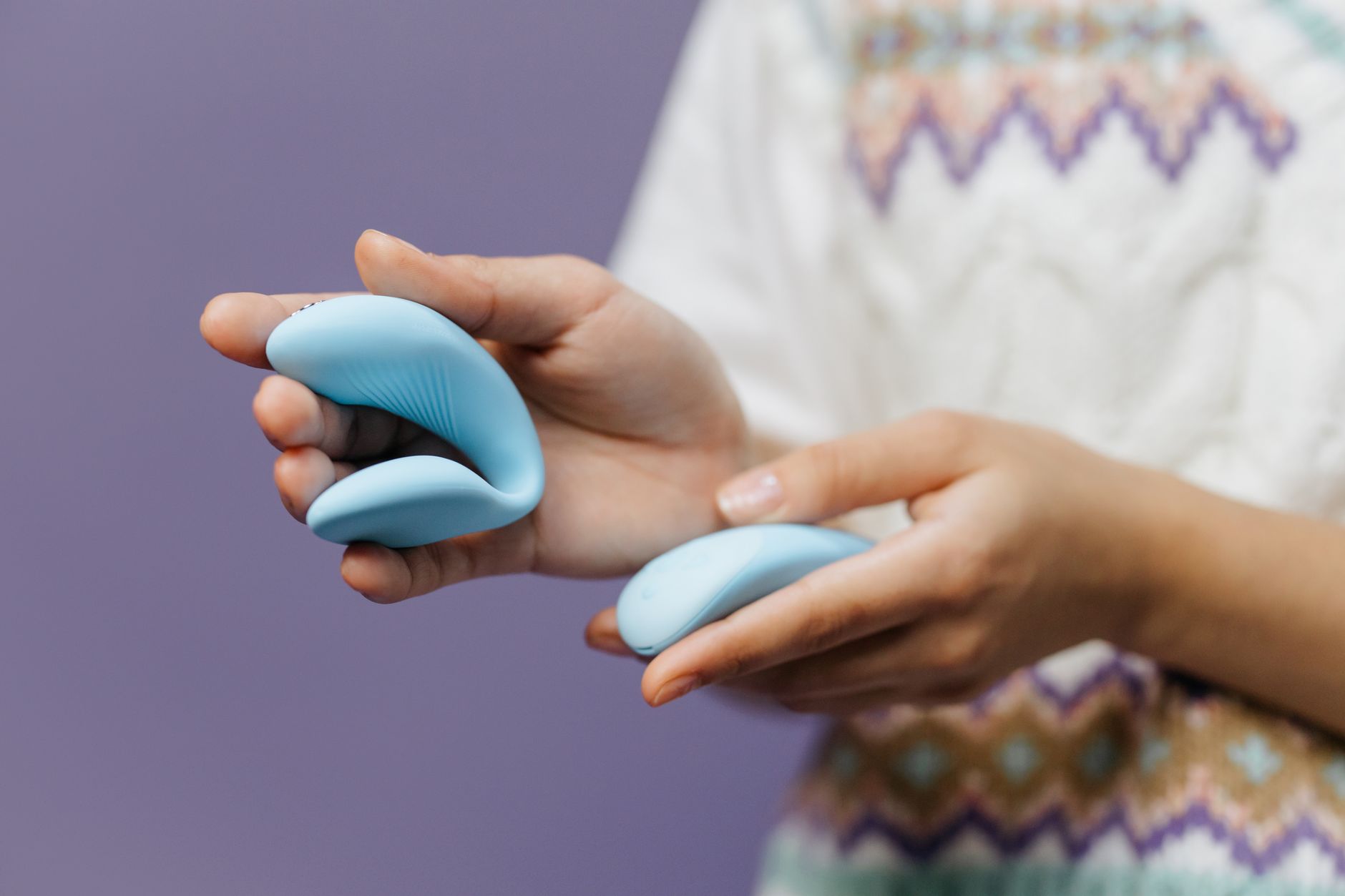 Close-up of hands gently holding a sleek blue clitoral vibrator against a purple background